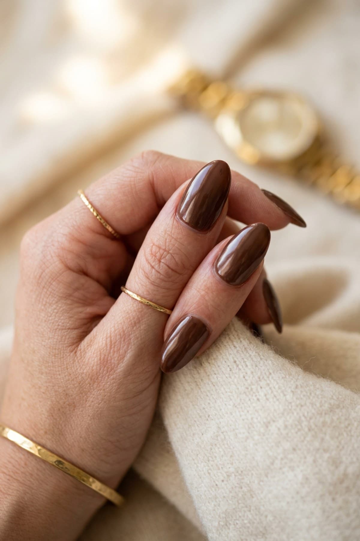 Glossy chocolate brown almond nails resting on a cream cashmere sweater with delicate gold rings and a gold watch